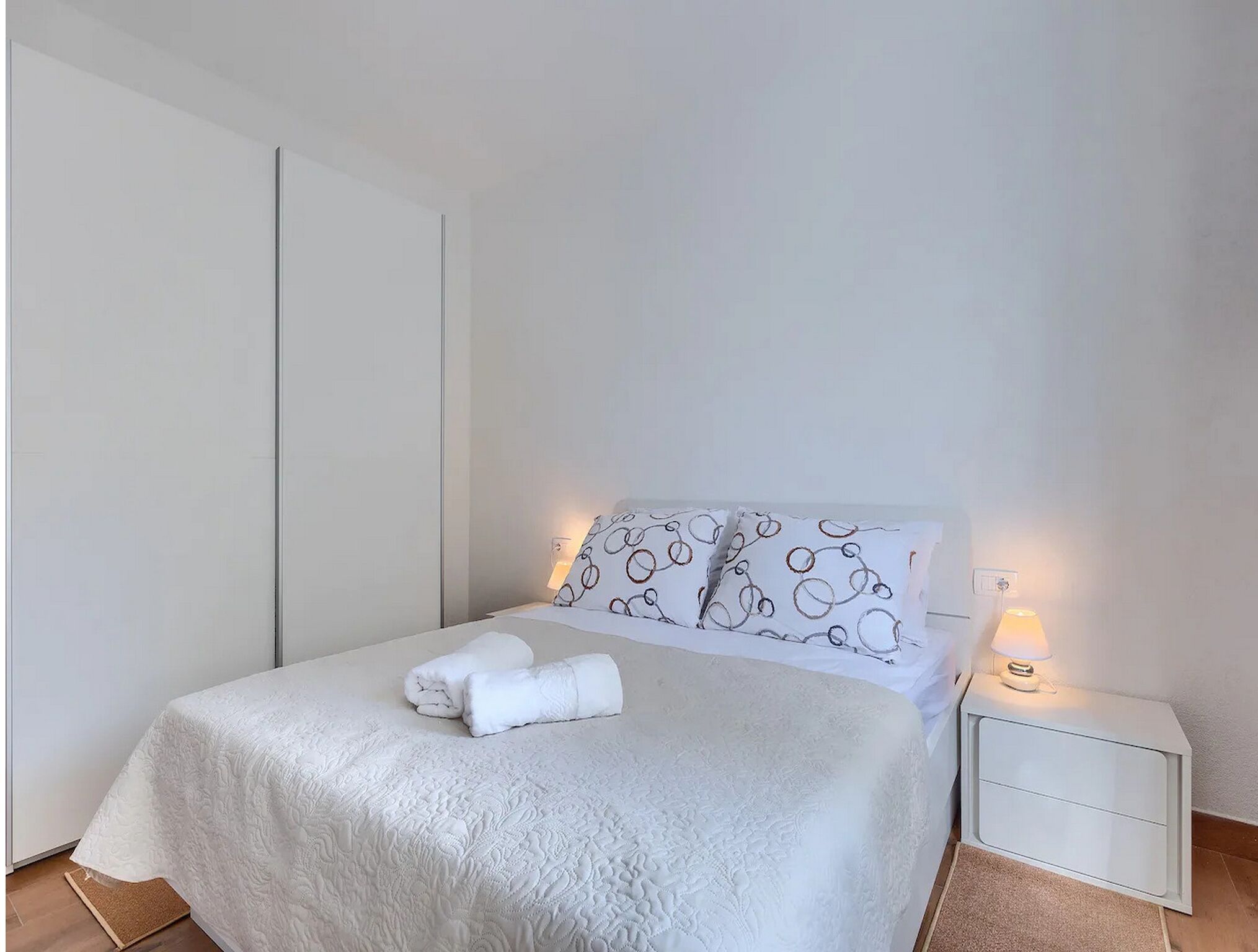 En pænt redt seng med et hvidt dynebetræk, to puder med et cirkulært mønster og et foldet håndklæde på sengen. Der er et sengebord med en lampe og en lille skuffe.