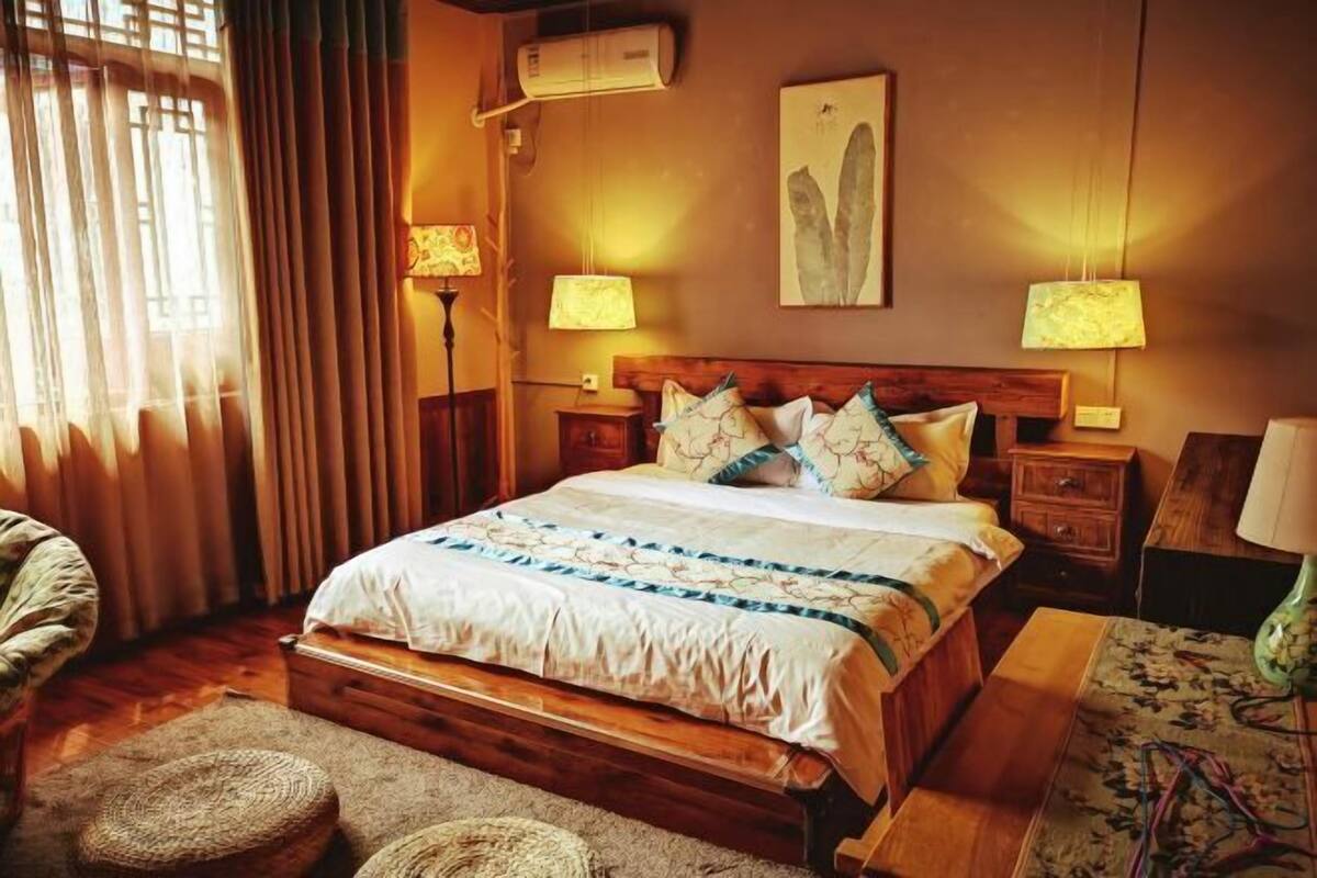 A bedroom with a wooden bed, two bedside lamps, a wall-mounted air conditioner, and a large window with curtains.