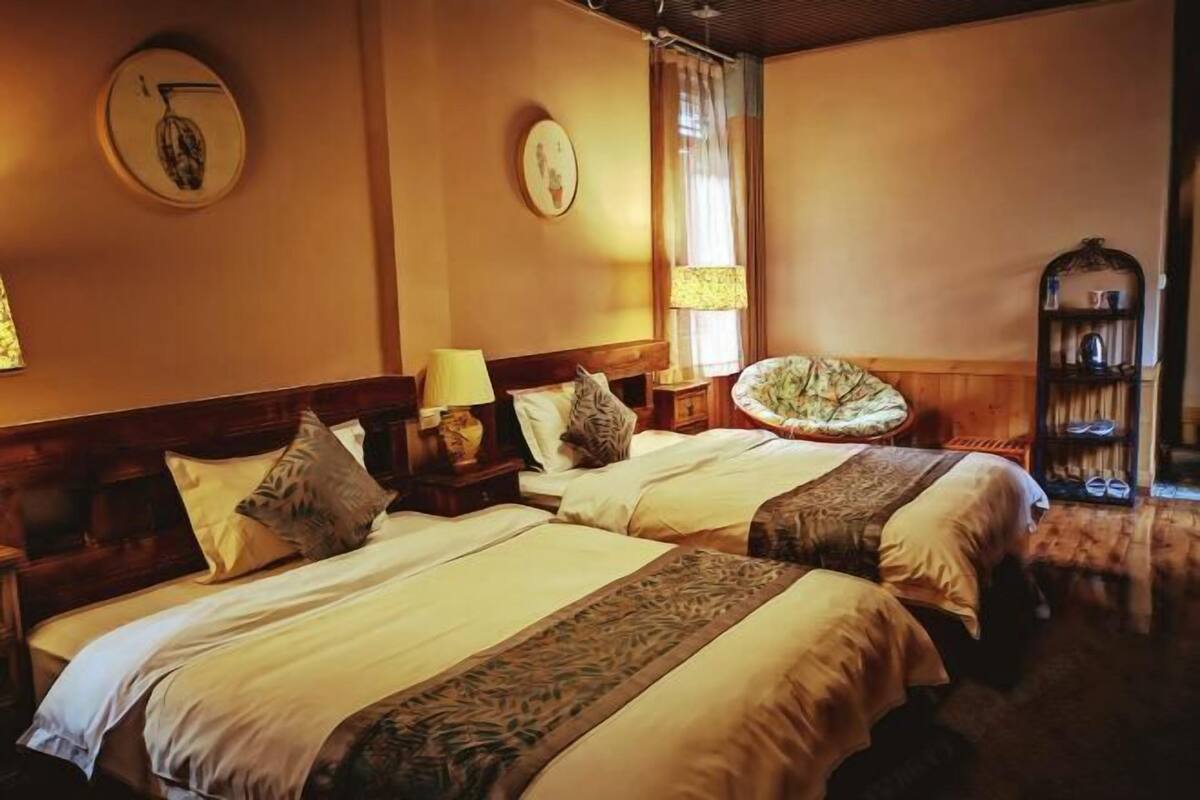 A hotel room with two beds, a wooden headboard, a small table with a lamp, a chair, and a shelf with shoes.