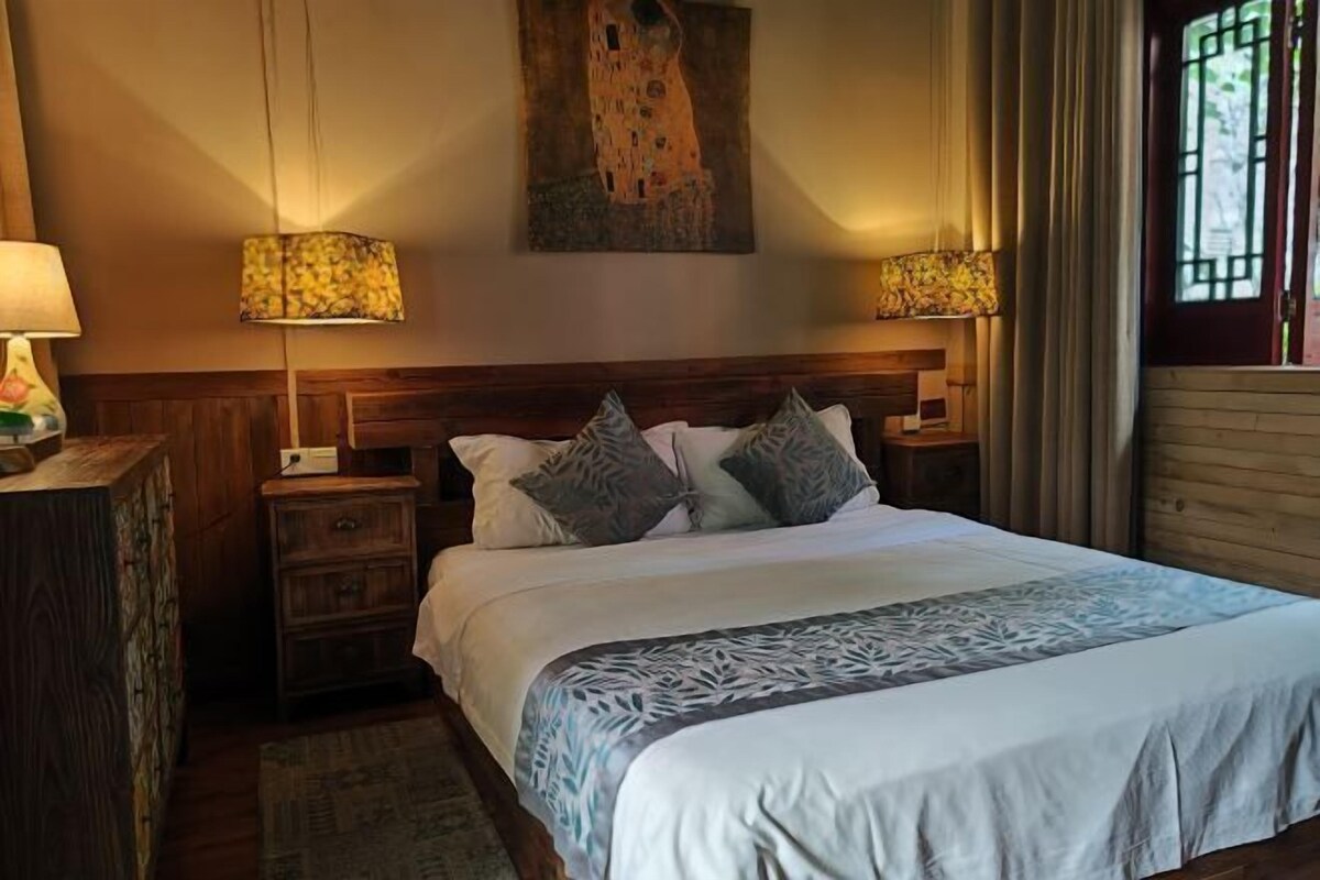 A neatly made bed with a patterned bedspread, two bedside lamps, a wooden headboard, and a painting above the bed.