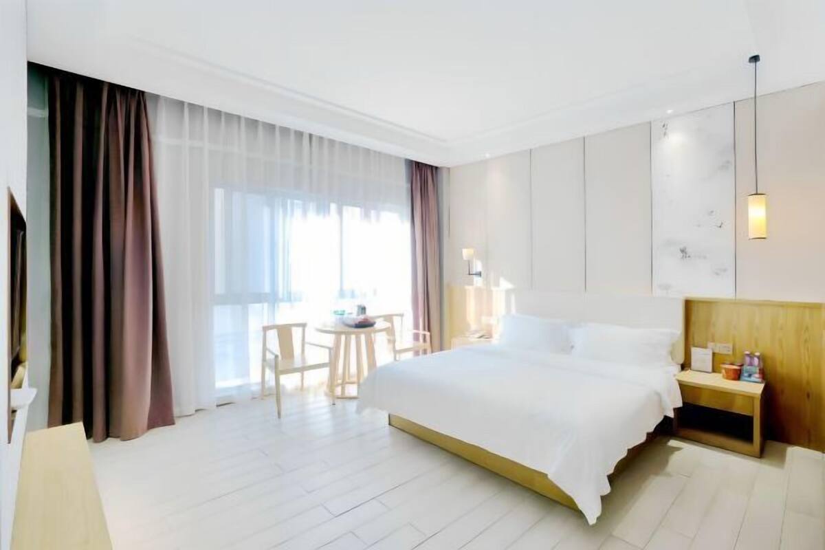 Habitación de hotel con una cama grande, una mesita de noche, un escritorio pequeño con una silla y una ventana con cortinas.