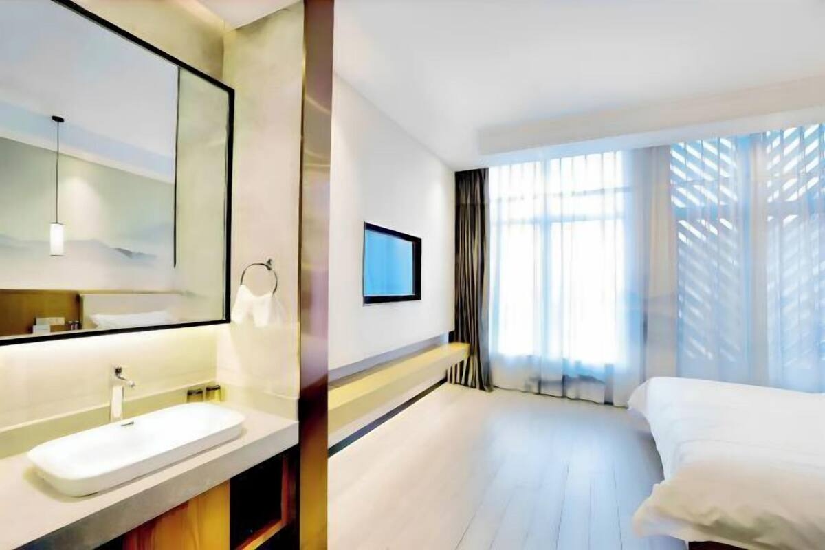 Una habitación de hotel moderna con una cama grande, un tocador empotrado con espejo, un televisor de pantalla plana en la pared y un amplio ventanal con cortinas translúcidas.