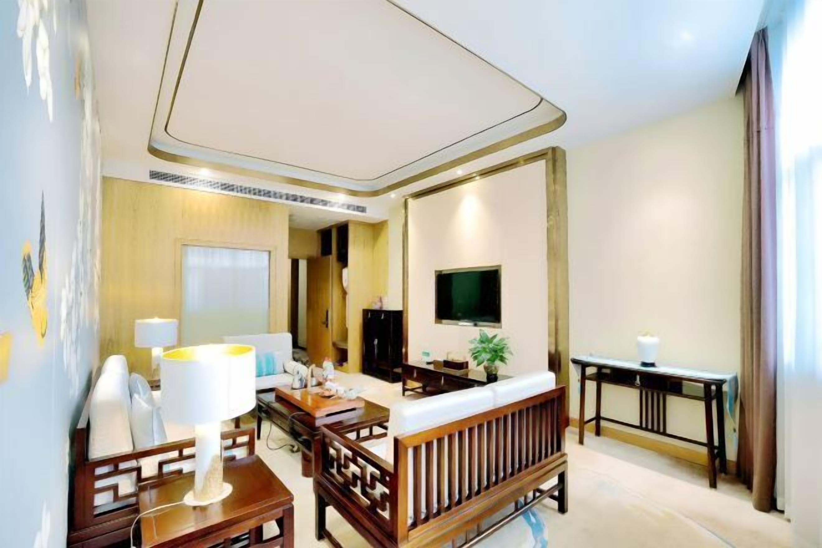 A hotel room with a wooden sofa, a TV mounted on the wall, a desk, and a lamp.