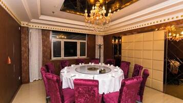 Una mesa de comedor redonda con mantel blanco, rodeada de sillas de terciopelo, un candelabro y un amplio ventanal con cortinas.