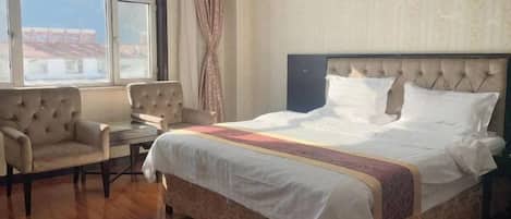 A hotel room with a large bed, a sitting area with two chairs, a small table, and a view of buildings through a large window.
