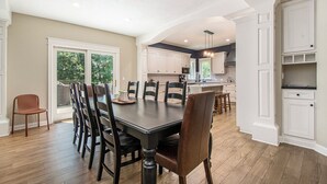 A spacious dining area with a large table and chairs, a kitchen in the background, and a view through the window.