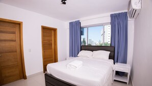 A bedroom with a bed, wooden doors, a window with curtains, and an air conditioning unit. - Lovely 2BR Spacious and Centric Apt. - Renty 501 (Santo Domingo)