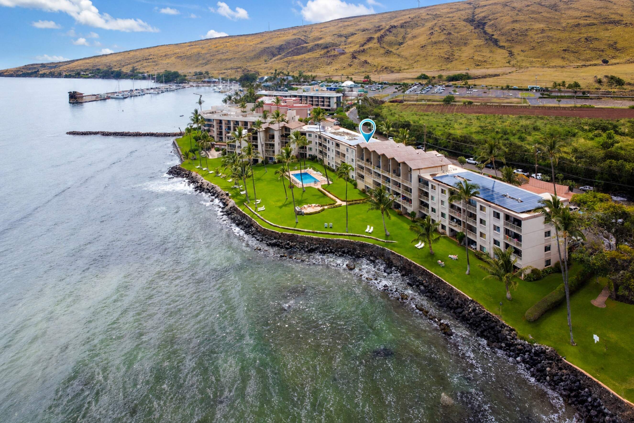 A coastal hotel with a swimming pool, multiple buildings, and a view of the sea and hills.