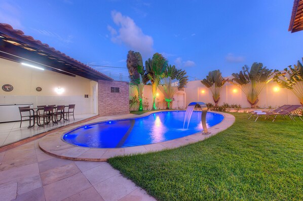 A swimming pool with a waterfall feature, surrounded by a well-maintained lawn and outdoor seating.