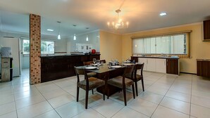 A spacious dining area with a large table set for six, surrounded by chairs. A kitchen area is visible in the background with modern appliances and a large window providing natural light. - Casa na Praia de Tabuba por Carpediem (Caucaia)