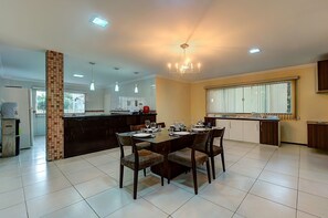 A spacious dining area with a large table set for six, surrounded by chairs. A kitchen area is visible in the background with modern appliances and a large window providing natural light. - Casa na Praia de Tabuba por Carpediem (Caucaia)