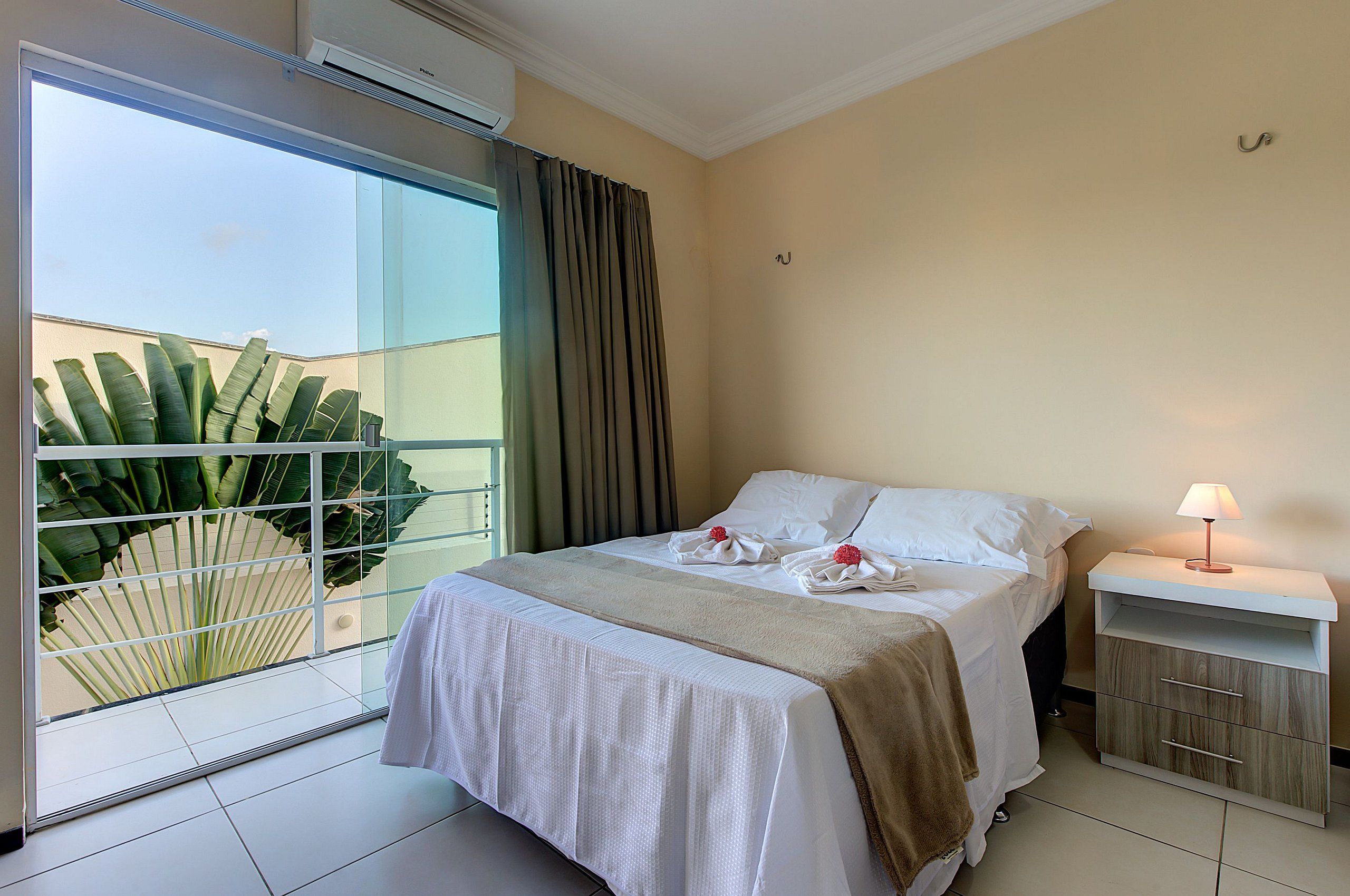 Una habitación de hotel con cama, mesita de noche y un amplio ventanal con cortinas.