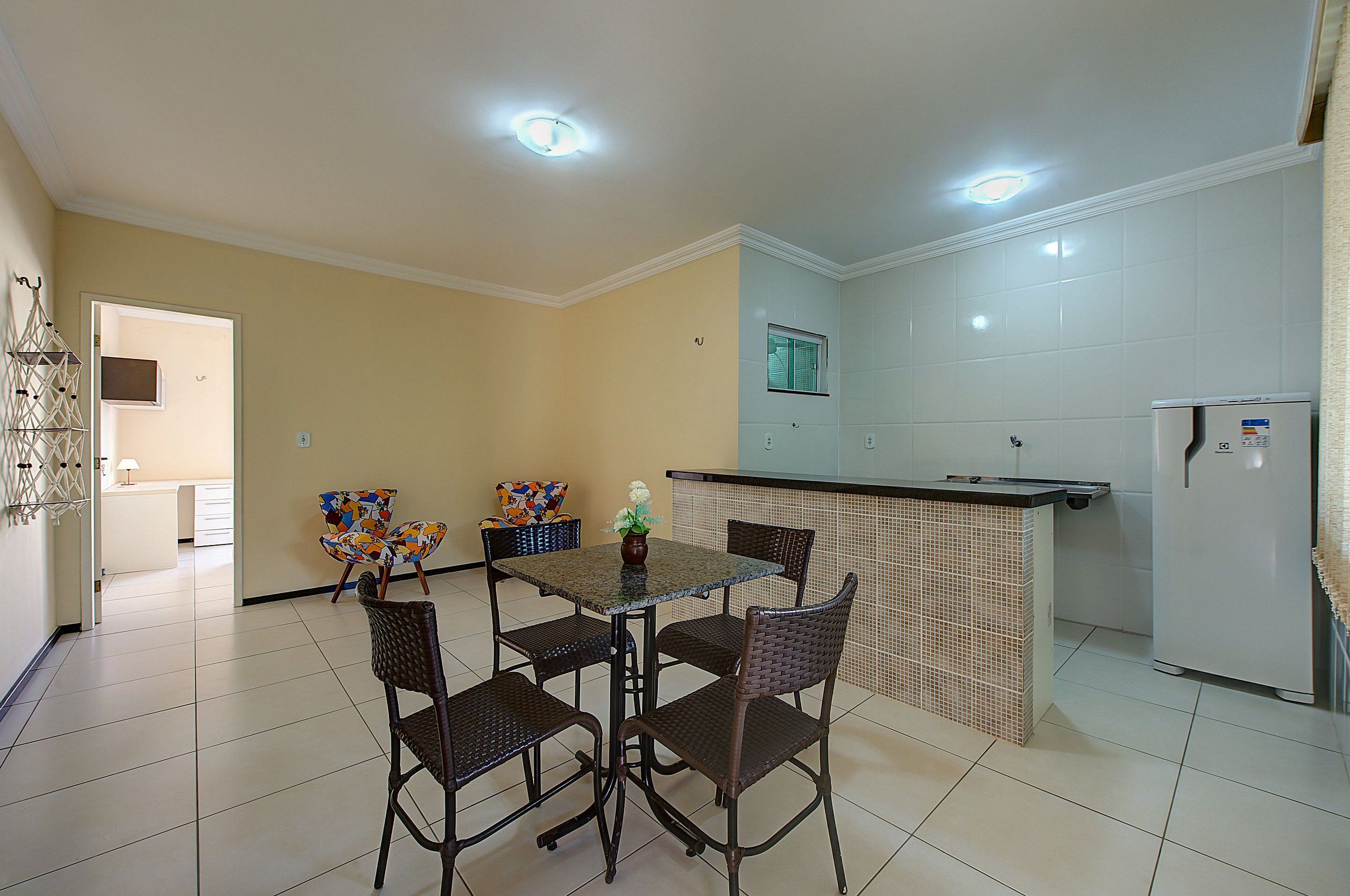 Un coin repas avec une table et quatre chaises, un petit espace cuisine équipé d’un réfrigérateur, et une fenêtre avec des stores.