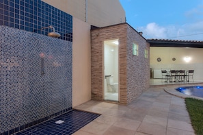 Um banheiro moderno ao ar livre com chuveiro, uma porta de vidro que dá para um vaso sanitário e uma área de piscina com mesa e cadeiras.