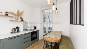 A modern kitchen with a white countertop, green cabinets, and a wooden dining table with a bench. There are pendant lights above the table, and a window with a view of the outside. - August-2523- (Cannes)