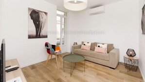 A modern living room with a beige sofa, a small round table, a colorful chair, a large elephant artwork on the wall, and a ceiling light fixture. - August-2523- (Cannes)