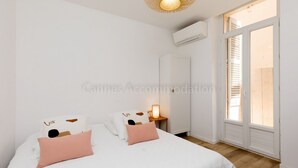 A bedroom with two beds, a bedside table with a lamp, an air conditioning unit, and a window with shutters. - August-2523- (Cannes)