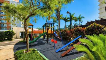 A playground with slides and climbing structures, surrounded by trees and a building in the background.