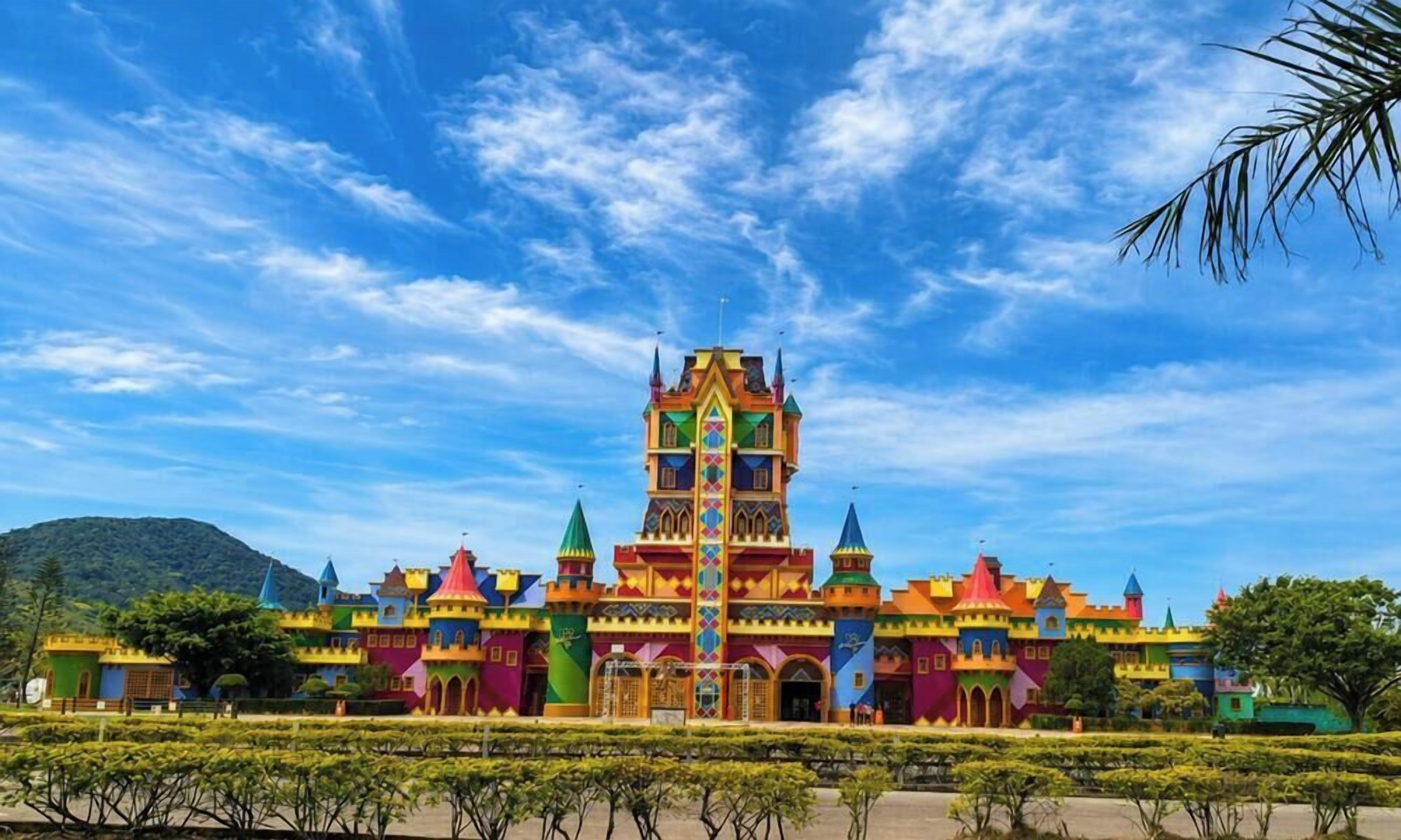 A colorful castle-like structure with multiple towers and a central spire, surrounded by well-maintained greenery.