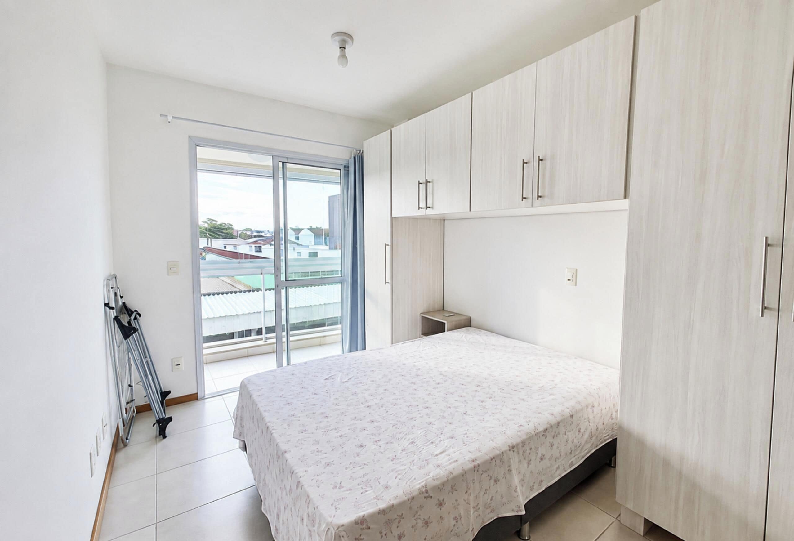 A bedroom with a bed, a wardrobe, a small bedside table, and a window with a view of buildings.