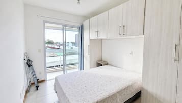 A bedroom with a bed, a wardrobe, a small bedside table, and a window with a view of buildings.