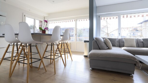 Un salon moderne avec un canapé d’angle gris, des chaises blanches et une table basse en bois.