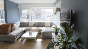 Un salon moderne avec un canapé d’angle, une table basse et une grande fenêtre donnant sur des immeubles et des arbres.
