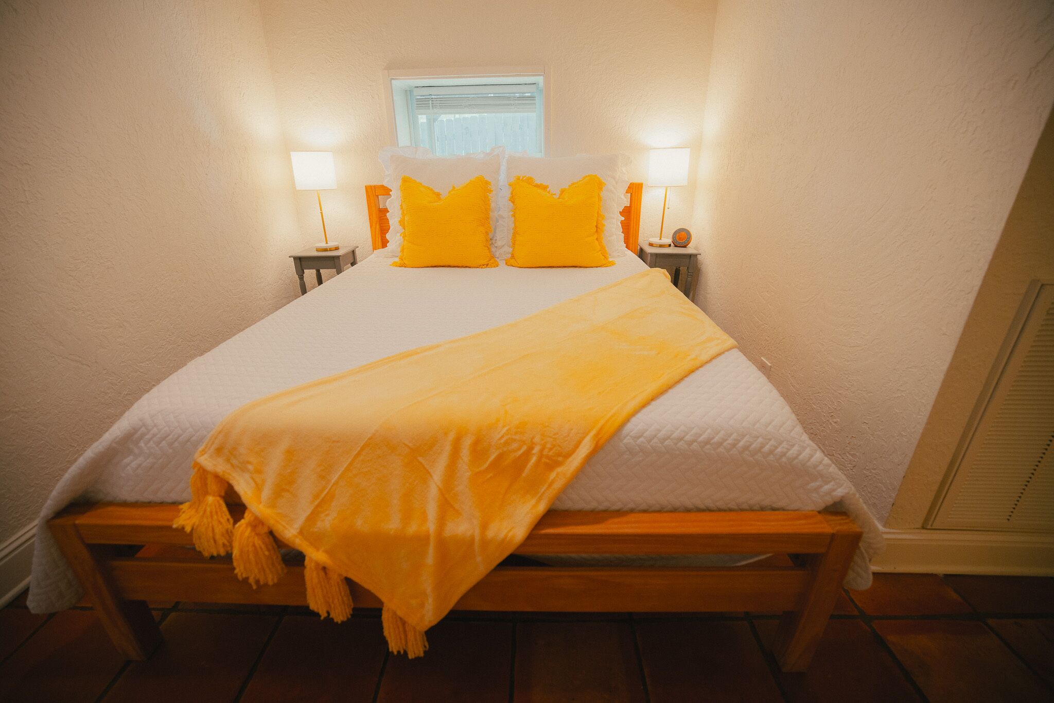Ein ordentlich bezogenes Bett mit weißer und gelber Bettdecke, zwei Nachttischlampen und ein Fenster darüber.