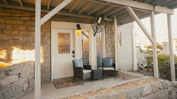 A covered porch with two wicker chairs and a table, a door with a number '5' sign, and a fan above the door.
