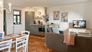 Una sala de estar moderna con un sofá gris, una mesa de comedor con sillas, una zona de cocina con electrodomésticos y un televisor sobre un mueble.