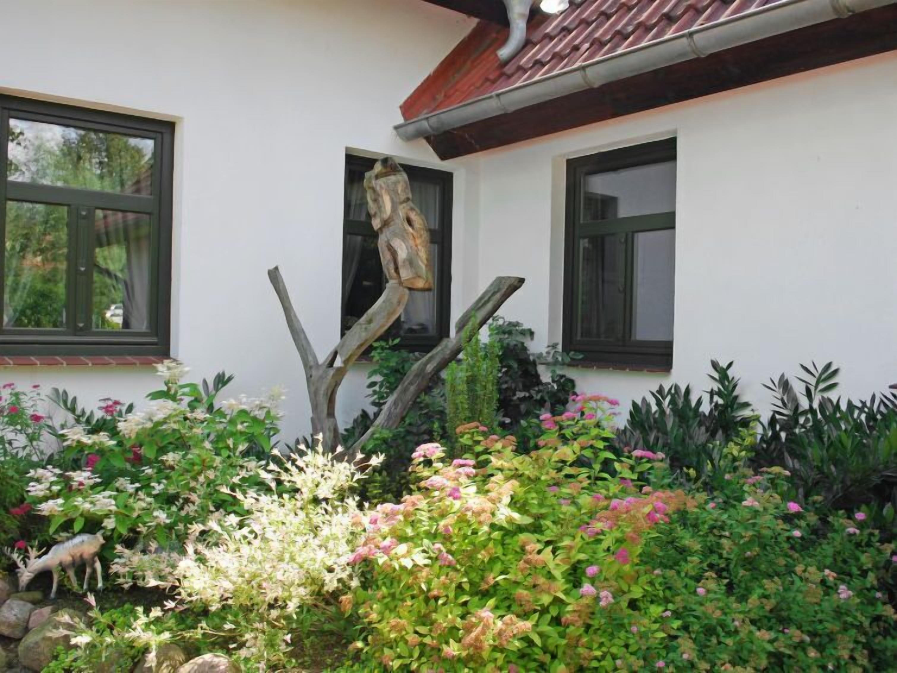 A garden with various plants and flowers in front of a building with two windows and a wooden sculpture.