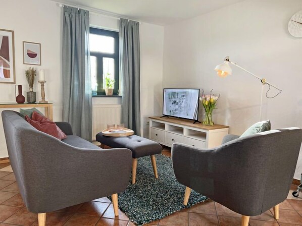 A modern living room with two grey sofas, a coffee table, a TV on a white cabinet, a floor lamp, and a window with curtains.