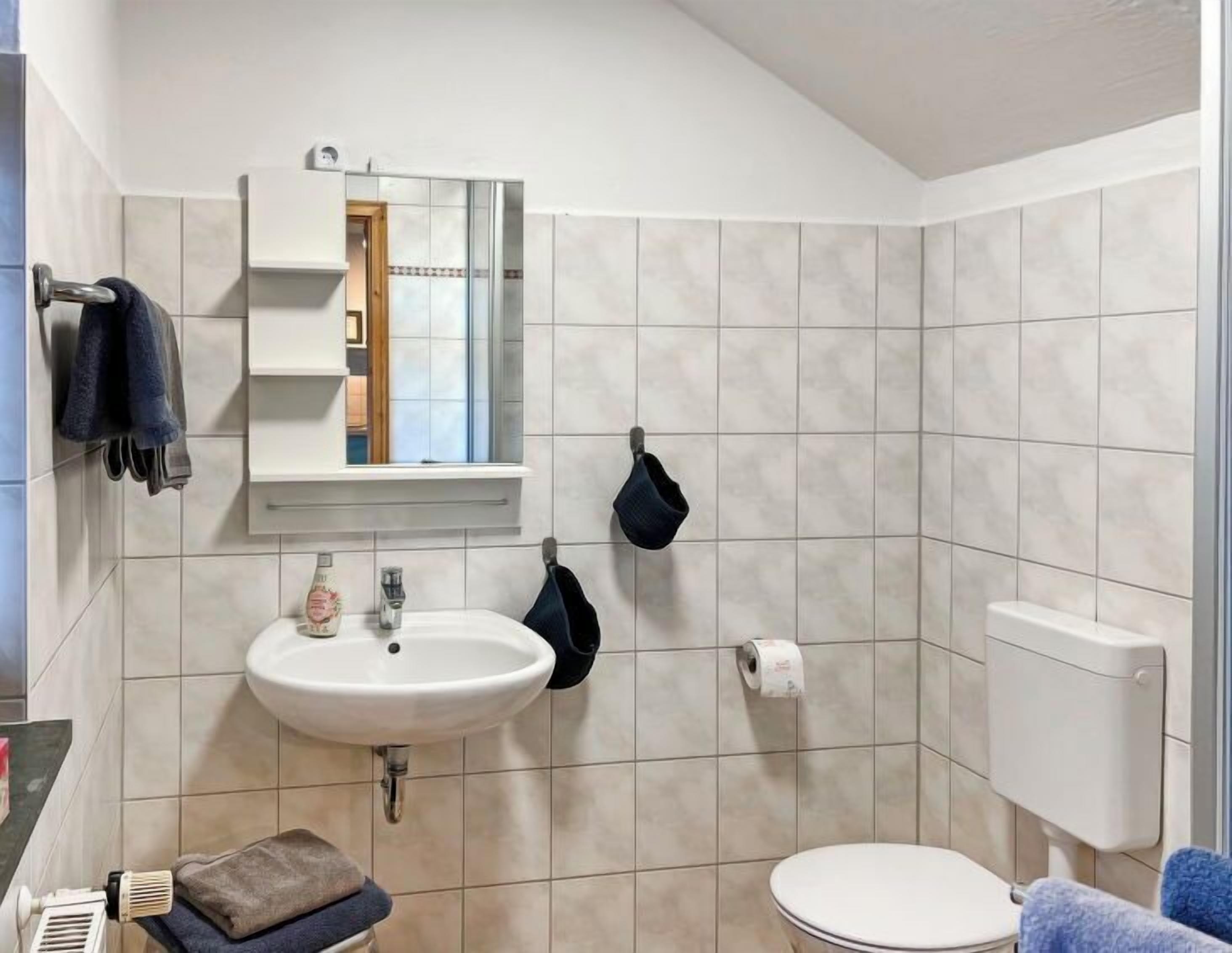 A bathroom with a white sink, mirror, and toilet. There is a towel rack with towels and a towel on the floor.