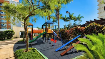 A playground with slides and climbing structures, surrounded by trees and a building in the background.
