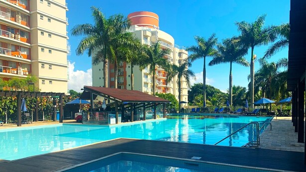 A swimming pool with lounge chairs and umbrellas, surrounded by palm trees and a multi-story building with balconies. - Nautilus Paradise in Penha-SC (Penha)