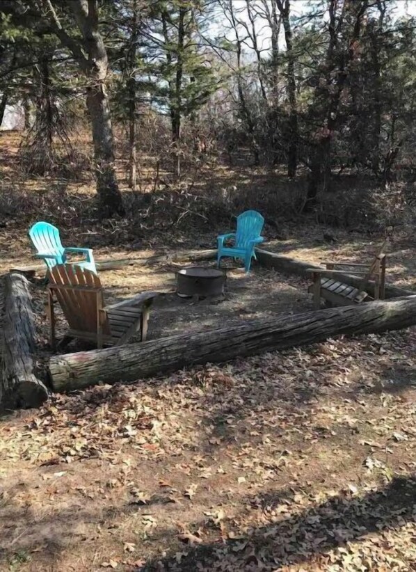 A forested area with two blue Adirondack chairs, a small fire pit, and a wooden bench.