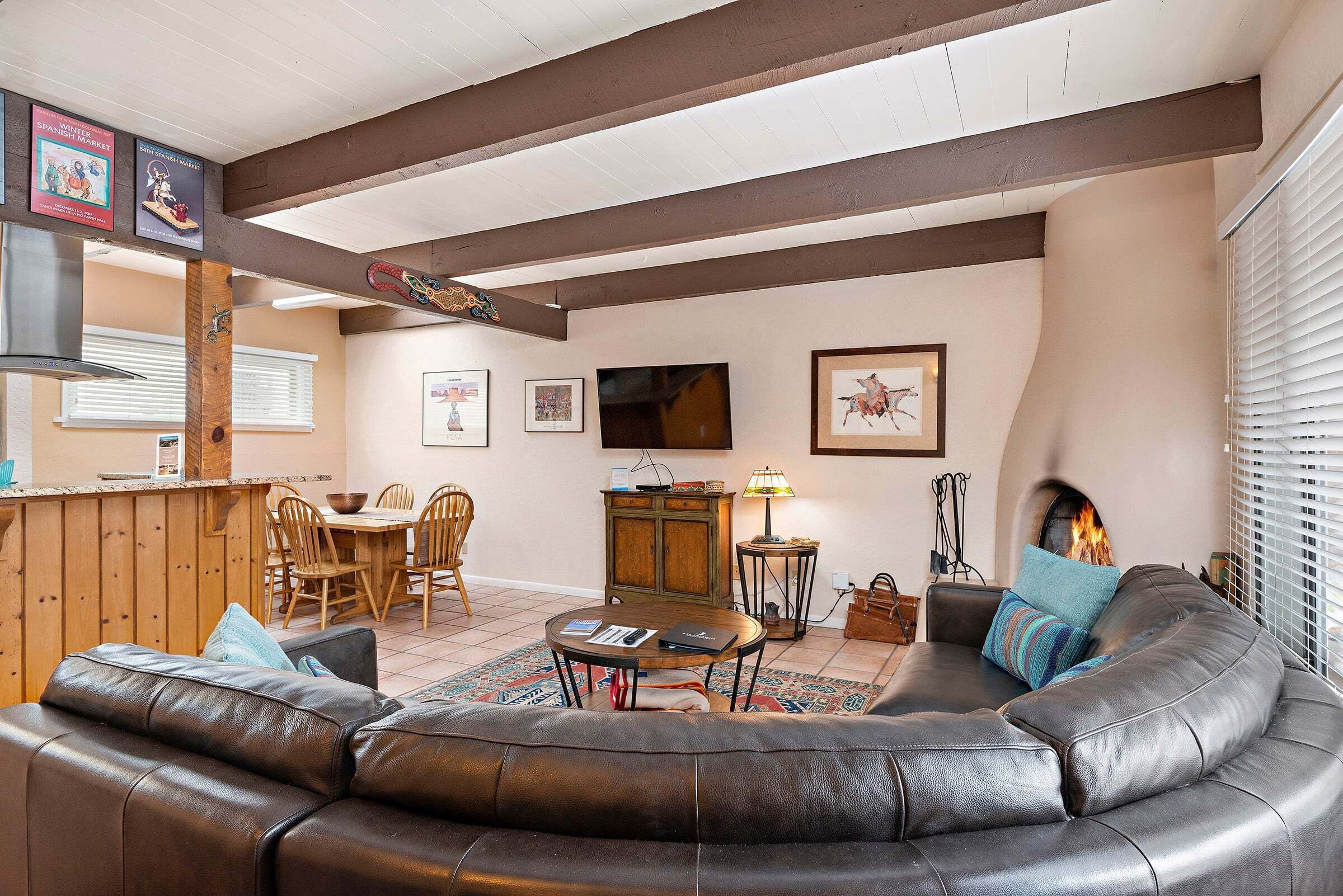 A living room with a leather sofa, wooden dining table, and chairs. There is a fireplace, a television, and framed artwork on the walls.