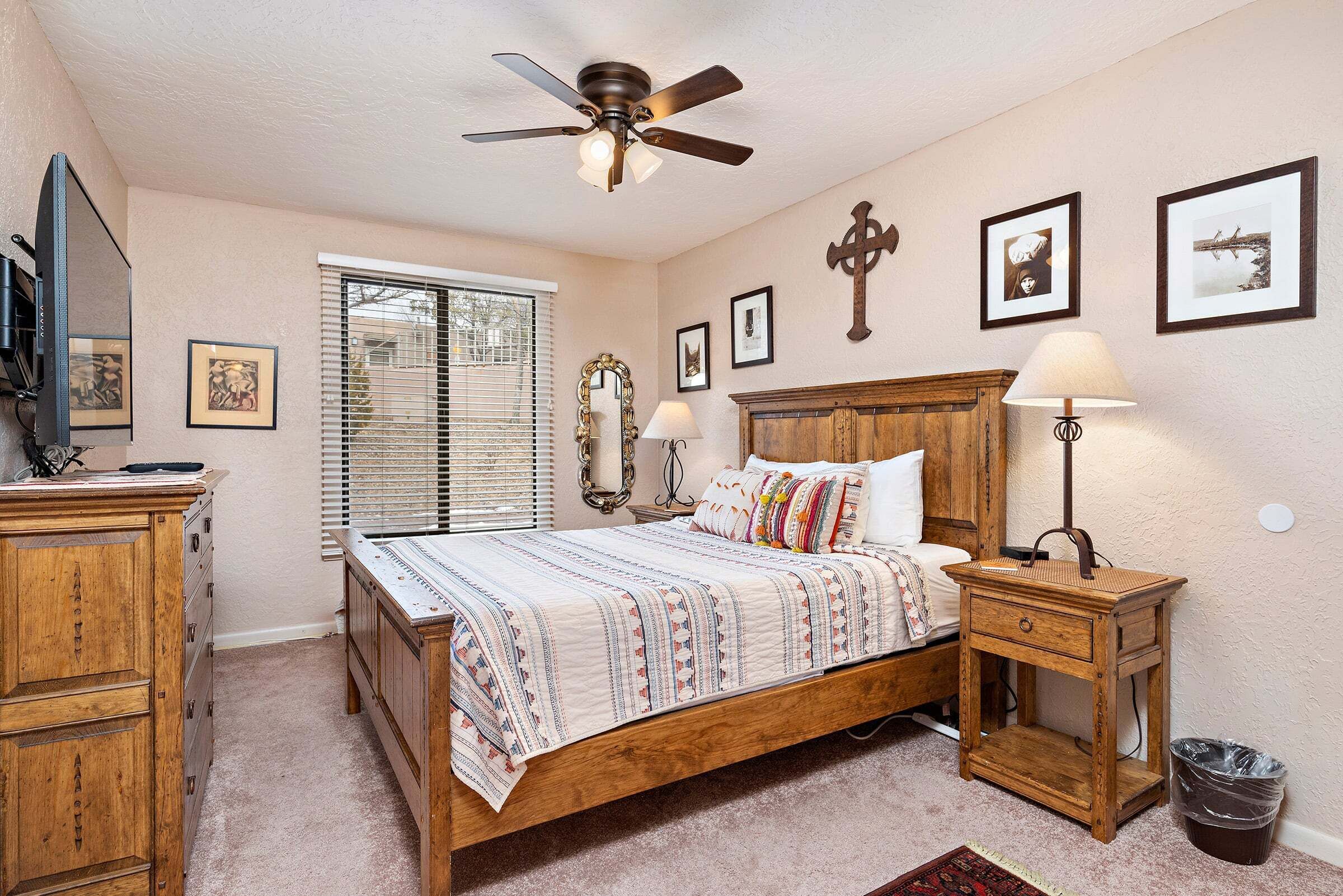 A bedroom with a wooden bed, a ceiling fan, a nightstand with a lamp, a dresser, and a window with blinds.