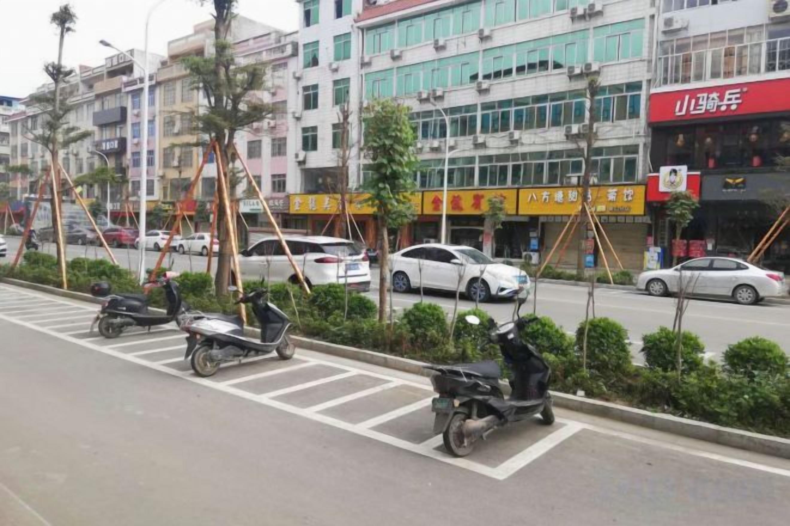 A street with parked scooters and cars, buildings with signage, and a pedestrian crossing.