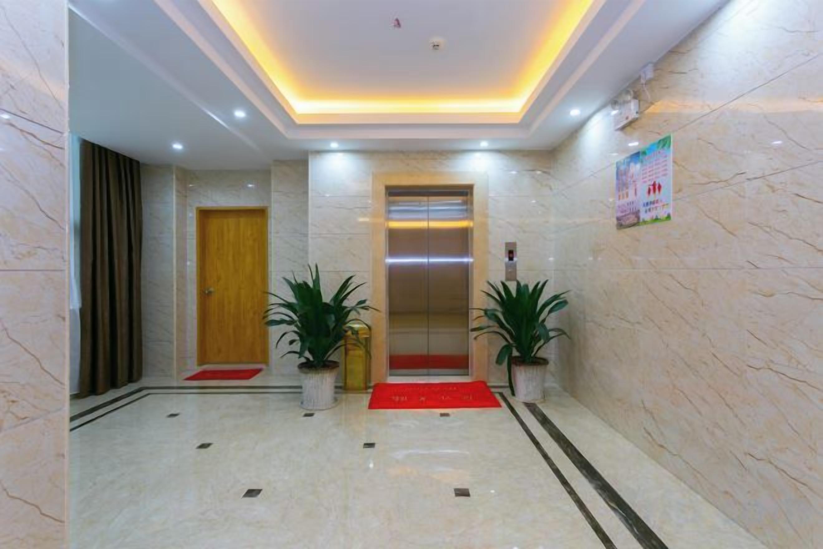 Vestíbulo de hotel con piso de mármol, alfombra roja, plantas en macetas, un ascensor y una puerta de madera.
