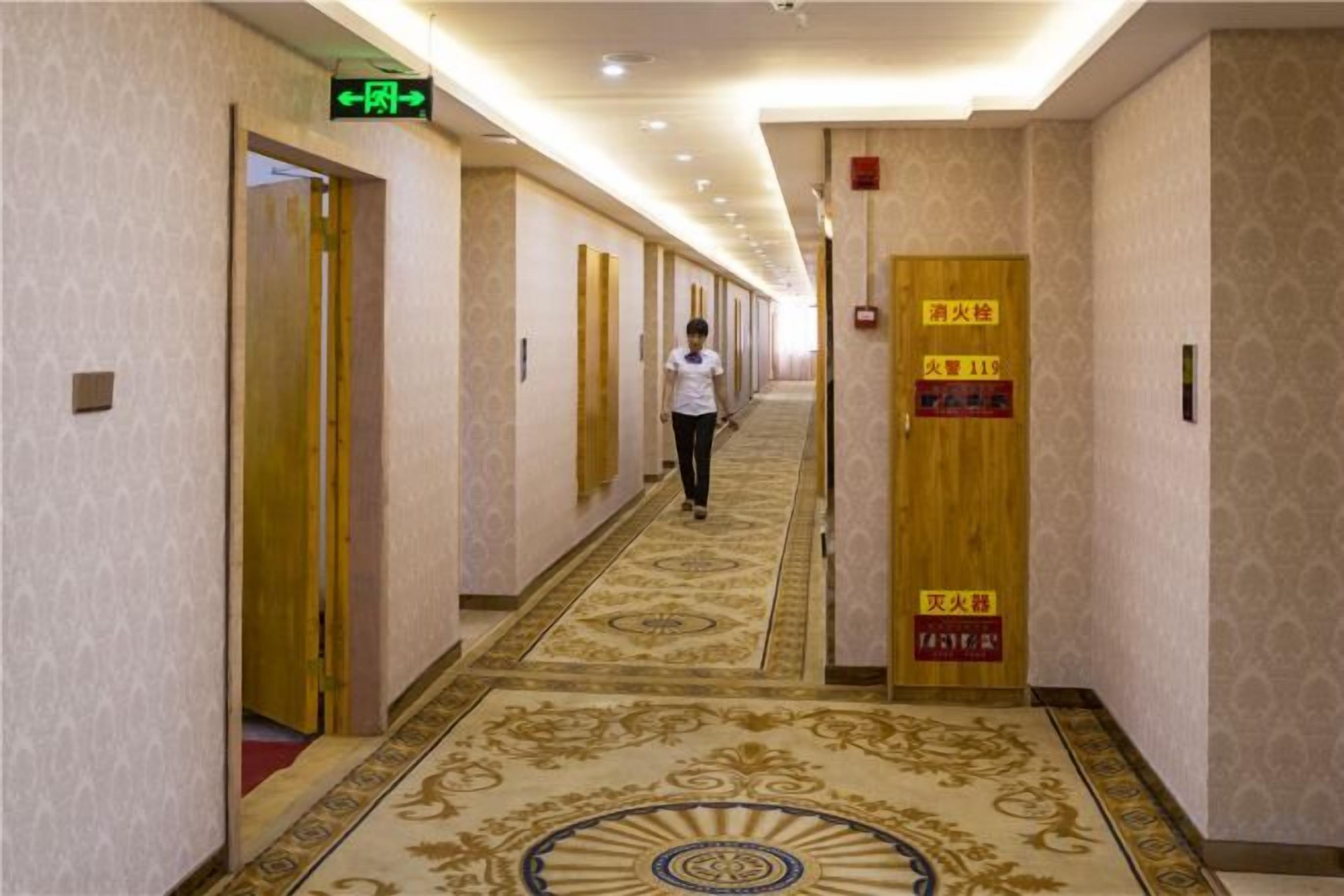 A hotel corridor with doors marked for rooms 11 and 12, a carpeted floor, and a woman walking down the hallway.