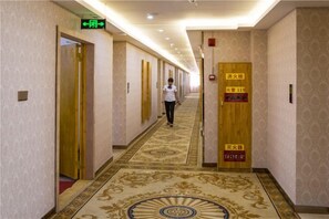 A hotel corridor with doors marked for rooms 11 and 12, a carpeted floor, and a woman walking down the hallway. - Wanxiang Hotel (Qinzhou Lingshan) (Lingshan)