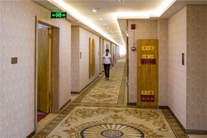 Un pasillo de hotel con puertas indicando las habitaciones 11 y 12, suelo alfombrado y una mujer caminando por el pasillo.