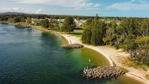 Ett kustnära område med fri sikt över en sandstrand, en lugn sjö och en rad hus i bakgrunden.