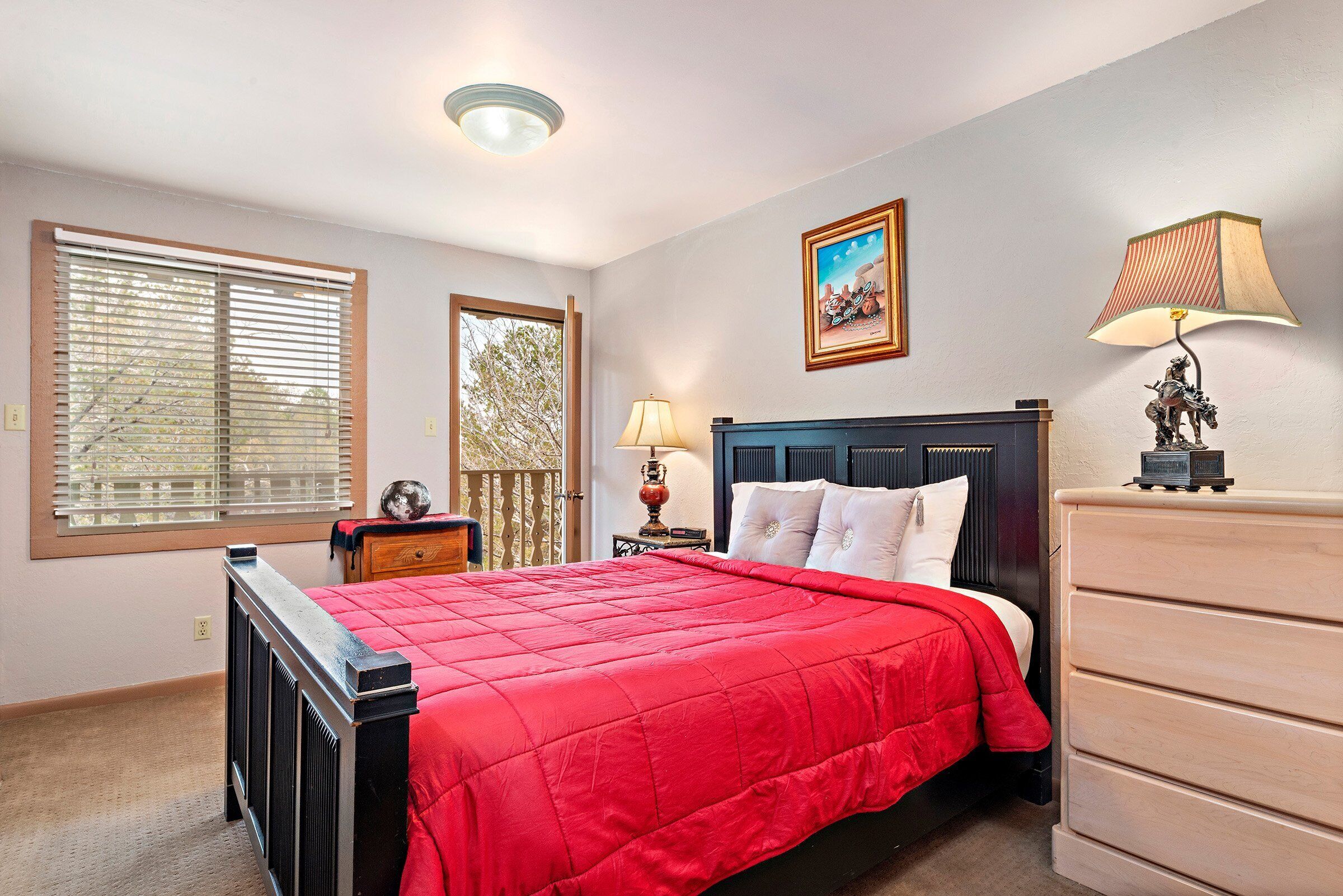 Een slaapkamer met een rode sprei, een houten dressoir, een nachtkastje met een lamp en een schilderij aan de muur.