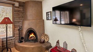 A living room with a fireplace, a television mounted on the wall, and a window with blinds. - Fort Marcy 43 | Walk to Plaza | Kiva Fireplace | Full Kitchen | Hot Tub | Pool | 2 Levels (Santa Fe)