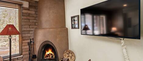 A living room with a fireplace, a television mounted on the wall, and a window with blinds.