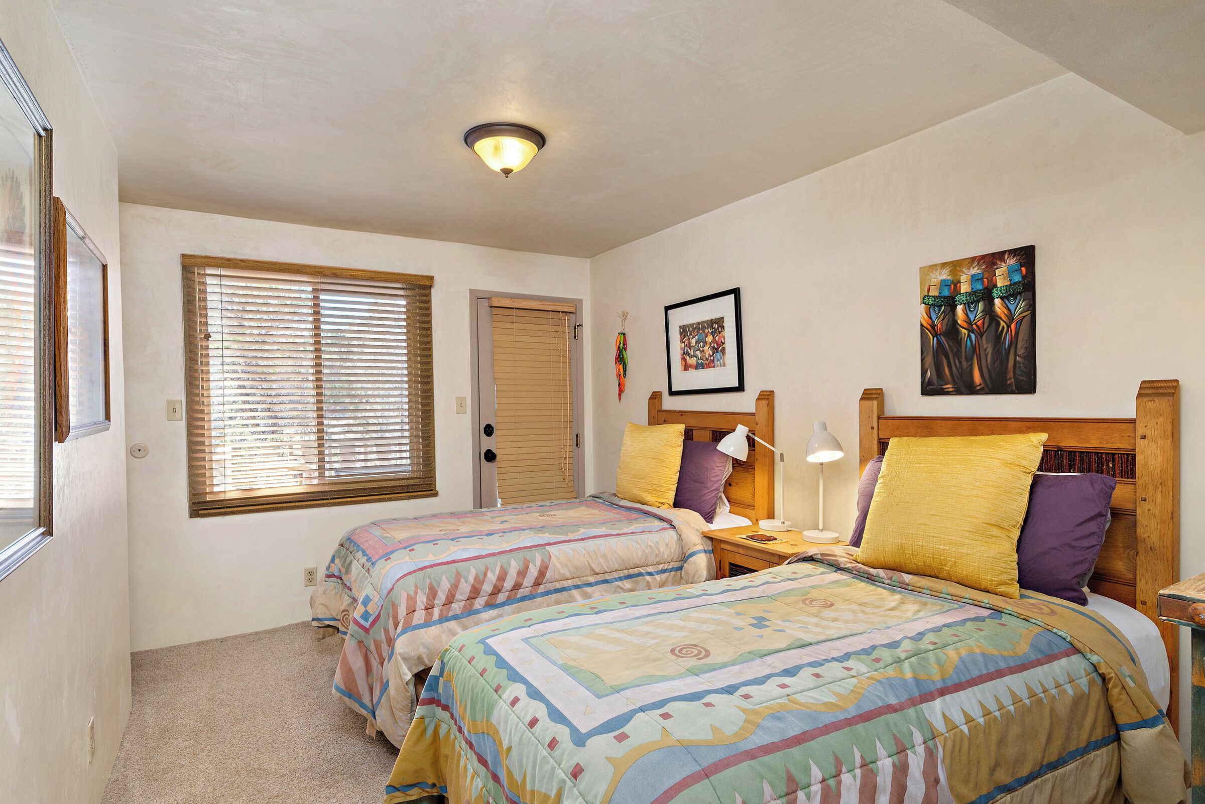 A bedroom with two beds, a window with blinds, a door, and a ceiling light.