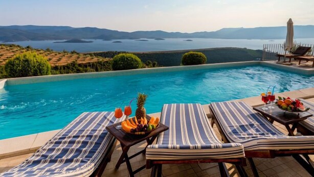 Un espace aménagé au bord de la piscine, avec des chaises longues et une table où sont disposés des fruits et des boissons, offrant une vue panoramique sur un paysage pittoresque composé de collines et d’une étendue d’eau.