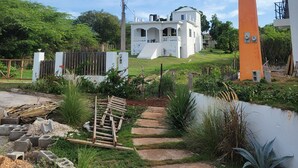 A white house with a large windmill in the foreground, surrounded by a garden with a stone pathway. - Dragonfly - House- Short walk to beach (Treasure Beach)
